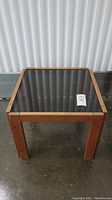 Top-front view of a square teak side table with a removable glass top, showing the full table against a plain background.