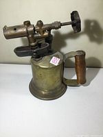 Front angle view of the old brass blow torch showing the cylindrical tank, wooden handle, valve knob, and nozzle assembly.