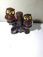 Front angled view of vintage owl porcelain salt and pepper set showing two brown owls with yellow eyes on a branch base.