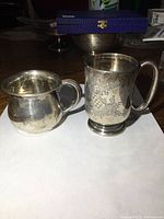 Pair of two sterling silver mugs shown side-by-side on white paper; one taller with floral engraving and pedestal base, the other shorter and rounder with smooth finish.