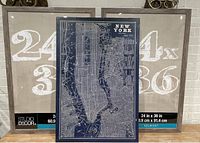 Two new Belmont 24x36 frames positioned side by side, with one frame featuring a large white printed label showing size '24 x 36' and brand 'Belmont'. A New York art poster with a dark blue detailed map of NYC is placed leaning against the frames.