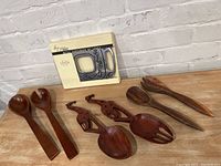 Full view of the lot showing Lenox Spyro metal trivet in box and six wooden serving utensils including giraffe-shaped spoons and salad servers.
