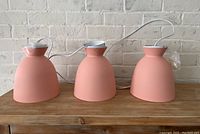 Three pink ceiling light fixtures placed side by side on a wooden surface against a white brick wall, showing their shape and finish.