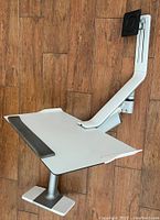 Side angle of the white computer monitor and keyboard/mouse stand on a wooden floor showing adjustable arm and tray.