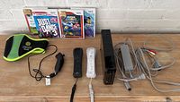 Photo shows the Nintendo Wii console with 3 controllers (2 Wii Remotes and 1 Nunchuk), sensor bar, AV cables, power adapter, and 4 game cases (Just Dance 3, Super Mario Galaxy, Go! Go! Mocha Squeak, Zumba Fitness). Small green wrist strap accessory also visible.
