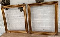 Two ornate gold wood frames leaning against white brick wall on wooden surface