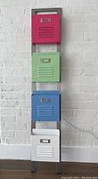 Front view of the metal locker magazine rack with pink, green, blue, and white compartments against a white brick wall.