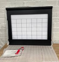 Front view of the black framed whiteboard calendar with a grid for days and mounting hardware on a cloth surface against white brick.
