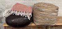 Photo showing two round poufs and two decorative throws on a white brick background. One pouf is dark brown pleather with wear, the other woven natural fiber. Throws folded on top of poufs, red and brown herringbone patterns visible.
