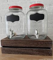 Front view of two glass beverage dispensers with red lids, chrome spigots, black chalkboard labels, and wooden display crate