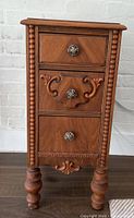 Front view of the vintage wooden side table displaying three drawers with intricate carved detailing and round decorative knobs; shows wear and surface marks.