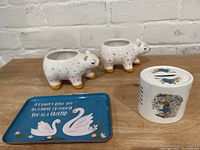 Two white ceramic bear vases and Peter Rabbit ceramic coin bank placed on surface with blue metal catchall tray showing swans and quote
