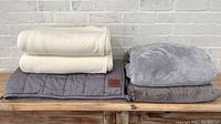 Four folded blankets and two blanket covers resting on a wooden surface in front of a brick wall. Gray blankets include weighted blankets and a plush cover in gray shades, and the covers in cream.
