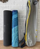 Two rolled yoga mats - one black and one blue with wave pattern - next to a gray and yellow yoga carrier bag with strap