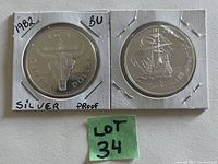 Two 1982 Canadian silver proof dollar coins in white paper holders with handwritten labels indicating 'BU' (Brilliant Uncirculated) and 'Proof Silver'. One coin shows a cow skull and 'Canada Dollar 1882-1982' text, the other a sailing ship.