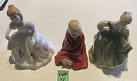 Three Royal Doulton figurines displayed together: 'April' in white dress, 'This Little Pig' child figurine wrapped in red shawl, and 'Fair Maiden' in green dress.