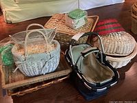 A collection of seven wicker baskets including a white wicker basket with fabric lining and handles. Several natural toned wicker baskets and one with red plaid fabric lining. Three metal framed baskets nested one inside another with fabric linings. Various sizes, some rectangular, some oval or round.