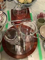 Two cheese boards with accessories displayed. The larger board has a glass dome cover, the smaller has three metal utensils. Catamount glass fat separator and two measuring cups are placed on boards.