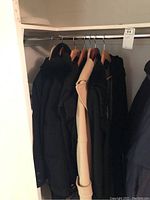 Six coats hanging on hangers in a closet, mostly dark colors with one light beige coat visible
