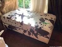 Rectangular cow hide covered ottoman with black and white spotted pattern shown in a room setting on wood flooring next to chairs.