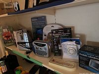 Photo showing various boxed and unpackaged gadgets including illuminated magnifier, labeler, Webster's spelling corrector, security timer, and other small electronic devices on a shelf.