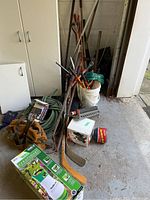 Assorted garden tools, hose, sprinkler and other items stacked on garage floor near cabinet.