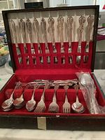Overall view of the silver plate flatware set in its original case showing knives arranged vertically in slots on back lid, forks and spoons placed horizontally in the lower section, and five serving spoons in the central area.