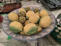 Top-down view of approximately twelve ceramic yellow lemons with green leaves arranged on blue and white patterned ceramic plate.