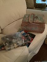Photo showing the layered woven sewing basket with floral covers next to a clear plastic case containing knitting needles, sewing notions, and knitting paper patterns underneath.