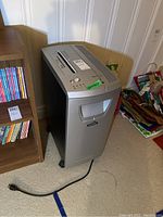 Photo of the Fellowes DM17Cs paper shredder placed on carpeted floor next to wooden bookshelf and bags, showing front and left side.