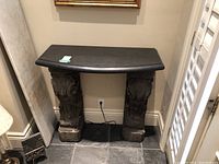 Side table with black marble top on two carved dark stone pedestal legs, with an extra marble slab leaning against the wall.