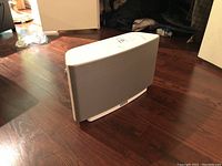 Side and angled view of white Sonos Play:5 wireless speaker with gray fabric grille, placed on wood floor.