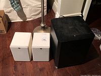 Two white RBH cube-shaped speakers and one black Samsung subwoofer placed on brown hardwood floor next to a lamp and white bed; speakers appear clean, subwoofer dusty.