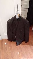 Dark double-breasted men's suit jacket hanging on hanger showing front view with tags attached.