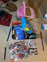 Large clear plastic bin filled with boxed games including Trivial Pursuit editions and LEGO, KNex sets spread on floor, with pink toy stroller visible.