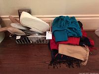 Winter apparel laid out on a surface including leather gloves in a basket and a stack of scarves and clothing items.