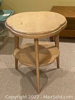 Photo of a three-legged wooden side table with scalloped round top and lower shelf, natural wood finish, standing on carpeted floor.