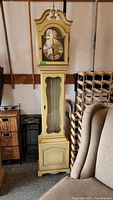 Full height image of grandfather clock showing cream yellow painted wood cabinet, bonnet with finial and glass door.