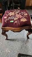 Full front view of the footstool showing wooden carved legs and floral needlework upholstery top.