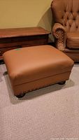 Large brown leather ottoman matched with chairs upholstered in similar brown leather with nailhead trim and tufted backs.