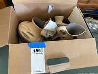 Open cardboard box containing three pottery pieces being sold as one lot. Items wrapped partially in newspaper and cloth for protection.