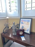 Overview of the lot items showing Asian figurine, metal bike sculpture, camera clock, and framed bicycle art print on a wooden table near window.