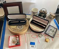 Full view of the lot showing cheese plate boxes, clocks, pens in cases, pocket watch, photo frame, and figurines.