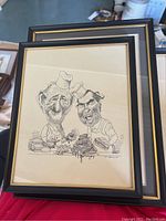 Photo showing one of two framed caricature prints of chefs preparing food, identifiable as political figures, in black frame with gold inner edge.
