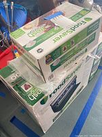 Boxes containing the FoodSaver vacuum sealing system and rolls of sealing bags shown stacked with other items in storage.