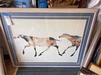 Framed poster depicting two horses in motion with visible signature and matting, wooden frame.