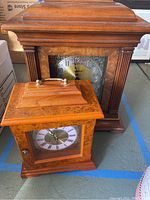 Two mantle clocks placed on the floor, one large dark wood Howard Miller clock behind a smaller lighter Gallery Collection clock
