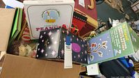 Box containing boxed 175 Peanuts plates set, a Charlie Brown Christmas book, and other Peanuts collectibles.