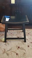 Front view of a black wooden stool with curved seat and four legs, placed on a patterned rug.