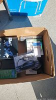 Box containing manuals, cables, Call ID device packaging, and some electronic accessories, photographed outdoors on concrete.